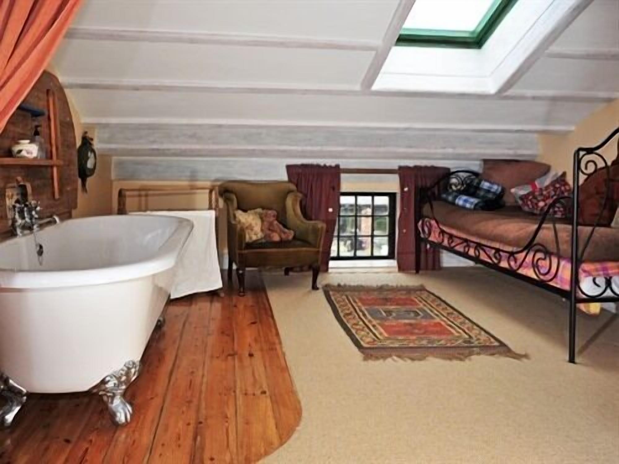 A bedroom with a large bathtub, a wooden floor, a window with curtains, and a bed with a patterned bedspread.