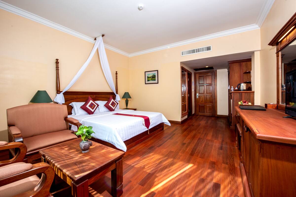 Quarto de hotel com cama, área de estar com poltrona e mesa, pequena mesa com planta e cozinha compacta.