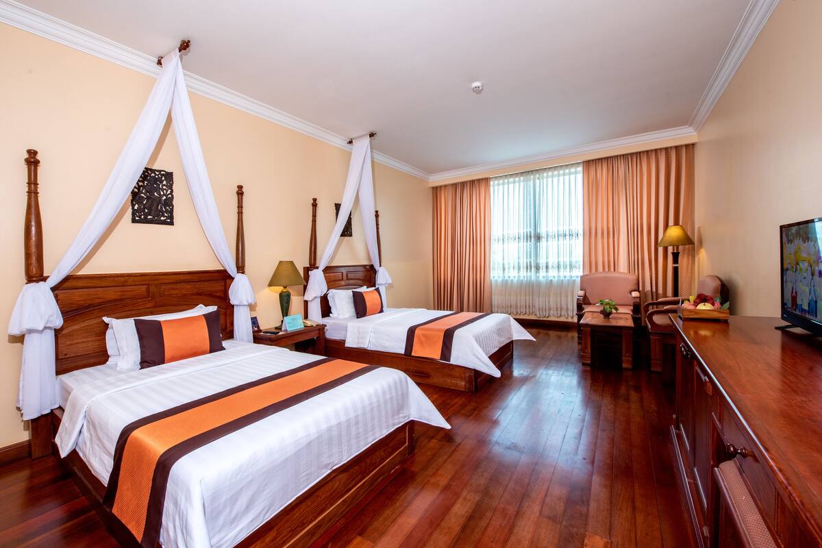 Quarto de hotel com duas camas, piso de madeira, televisão e uma mesa com abajur.
