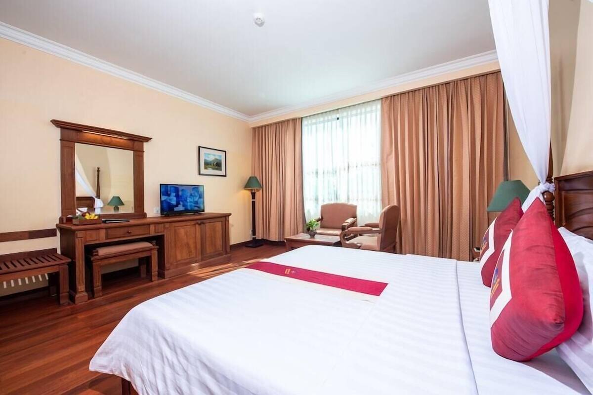Quarto de hotel com uma cama grande, uma escrivaninha com cadeira, televisão e janela com cortinas.