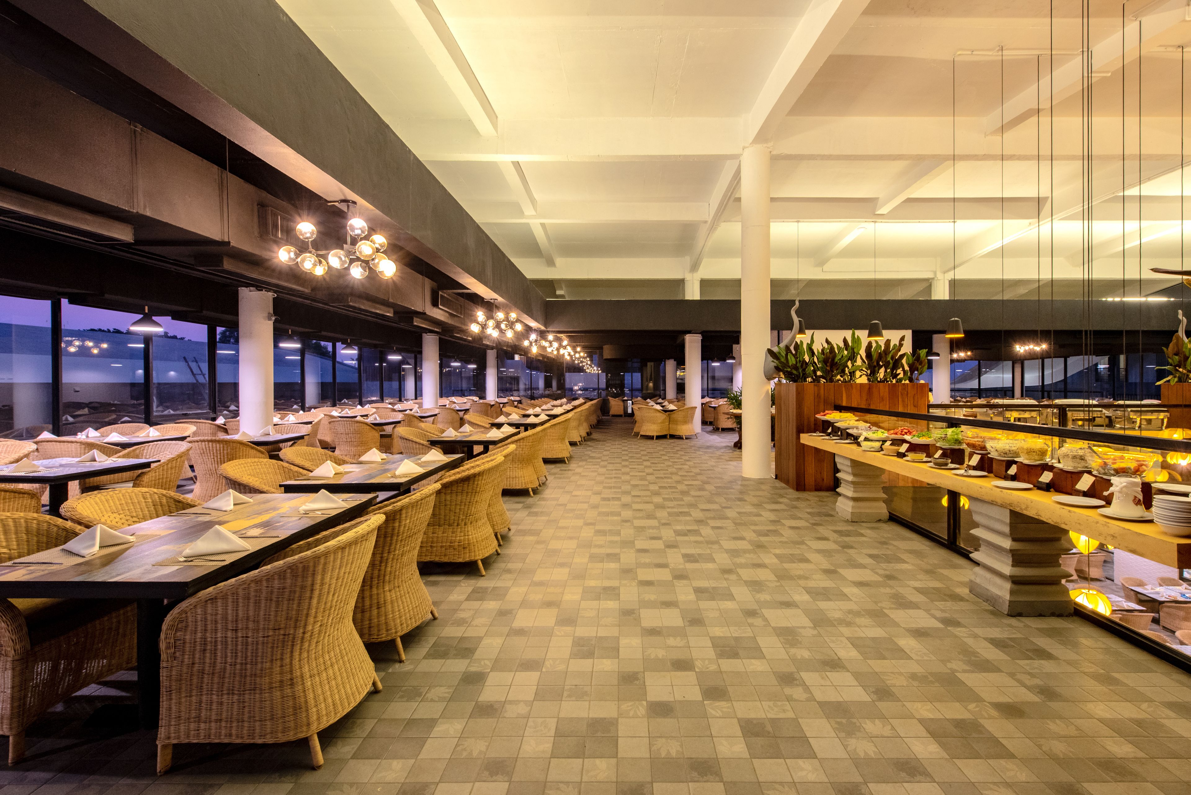 Un vaste espace dédié à la restauration, avec de longues tables et des chaises en osier, un buffet proposant une variété de plats, et de grandes fenêtres offrant une vue sur le paysage urbain.