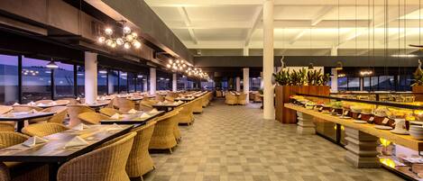 A spacious dining area with long tables and wicker chairs, a buffet counter with food, and large windows offering a view of the cityscape.