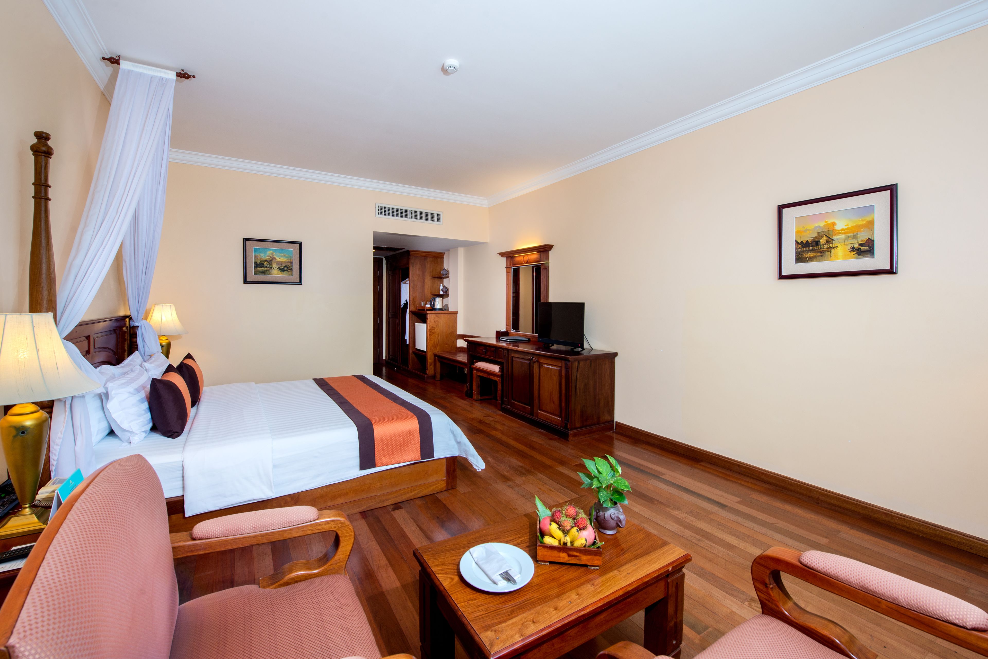 A hotel room with a bed, a desk, a television, and a dining table with fruit and a plant.