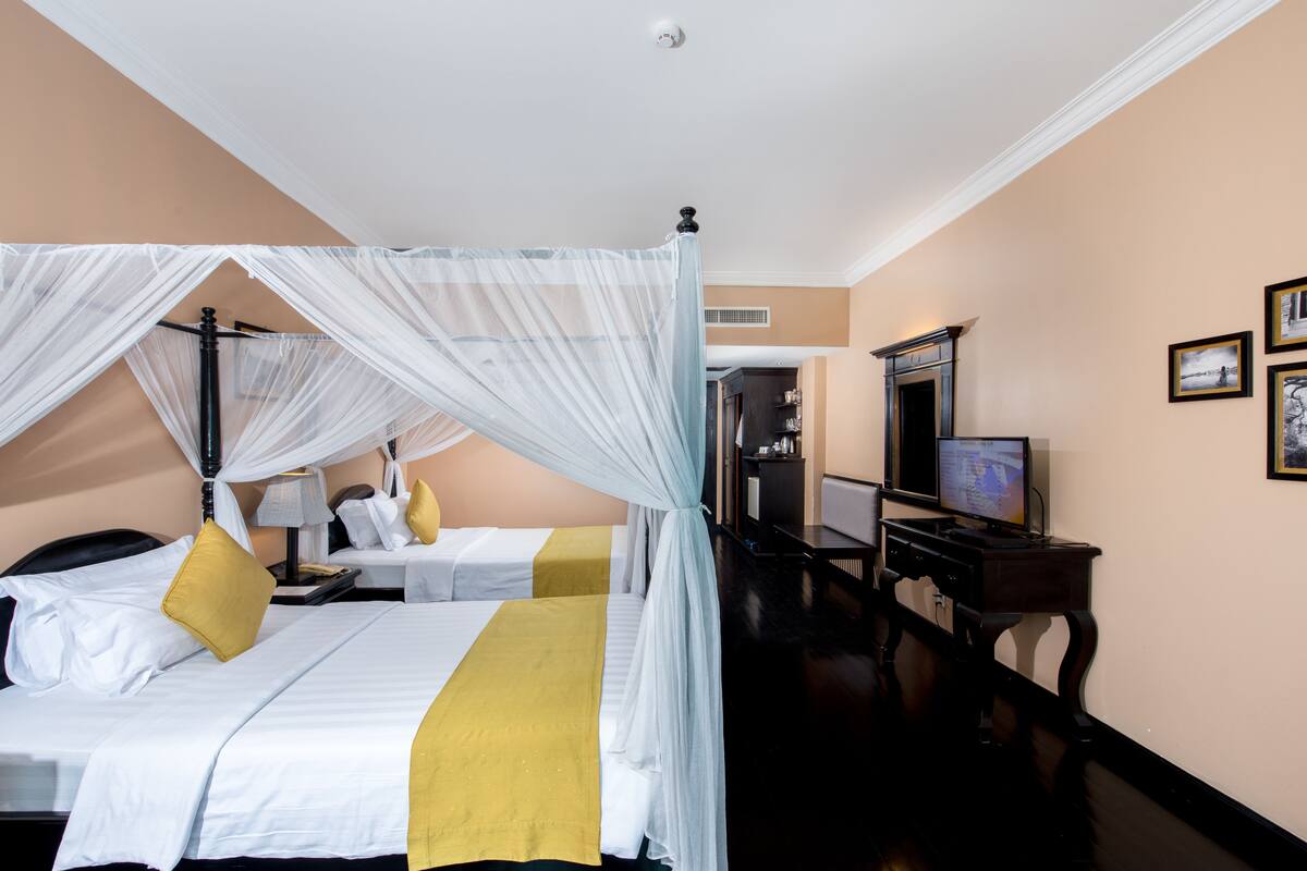 Quarto de hotel com duas camas, cada uma com dossel, uma televisão, uma escrivaninha e um espelho.