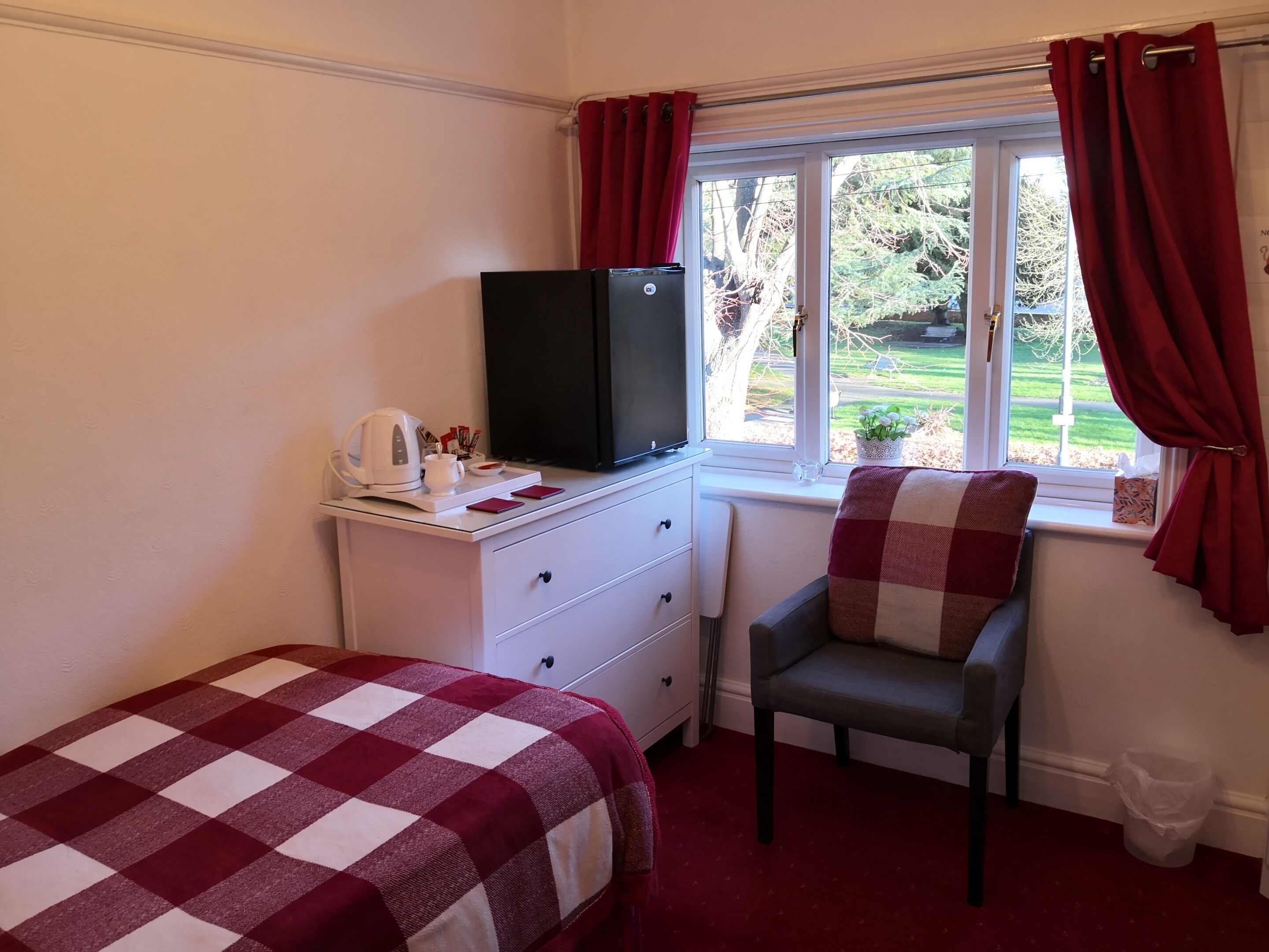 A hotel room with two beds, a desk with a kettle, a television, and a window with red curtains.