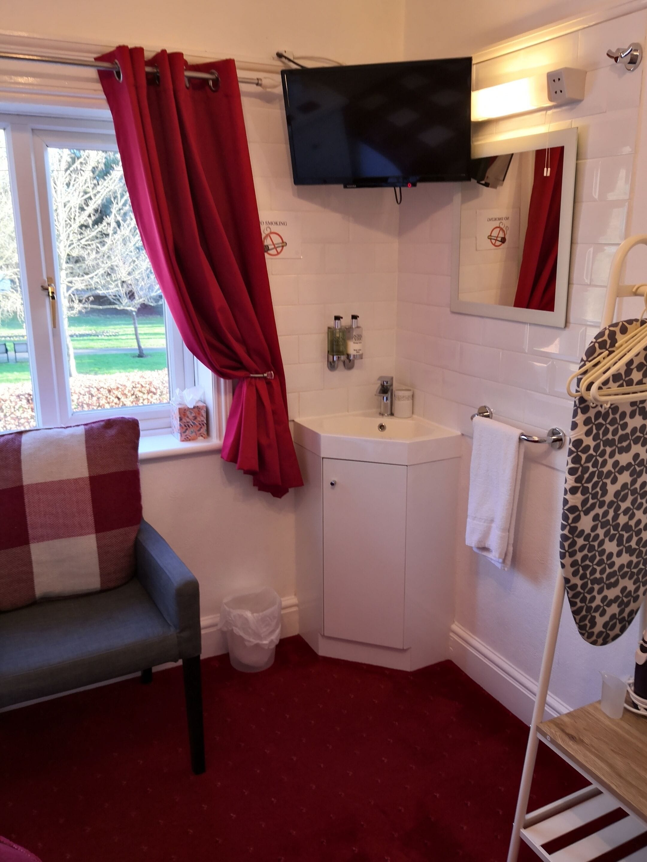 A bathroom with a white sink, a mirror, a television mounted above the sink, and a window with red curtains.