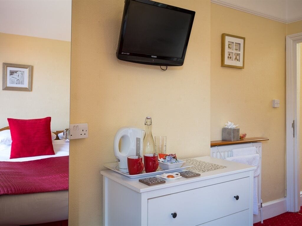 A hotel room with a bed, a bedside table with a TV, a red pillow, a white bedside table, and a framed picture on the wall.