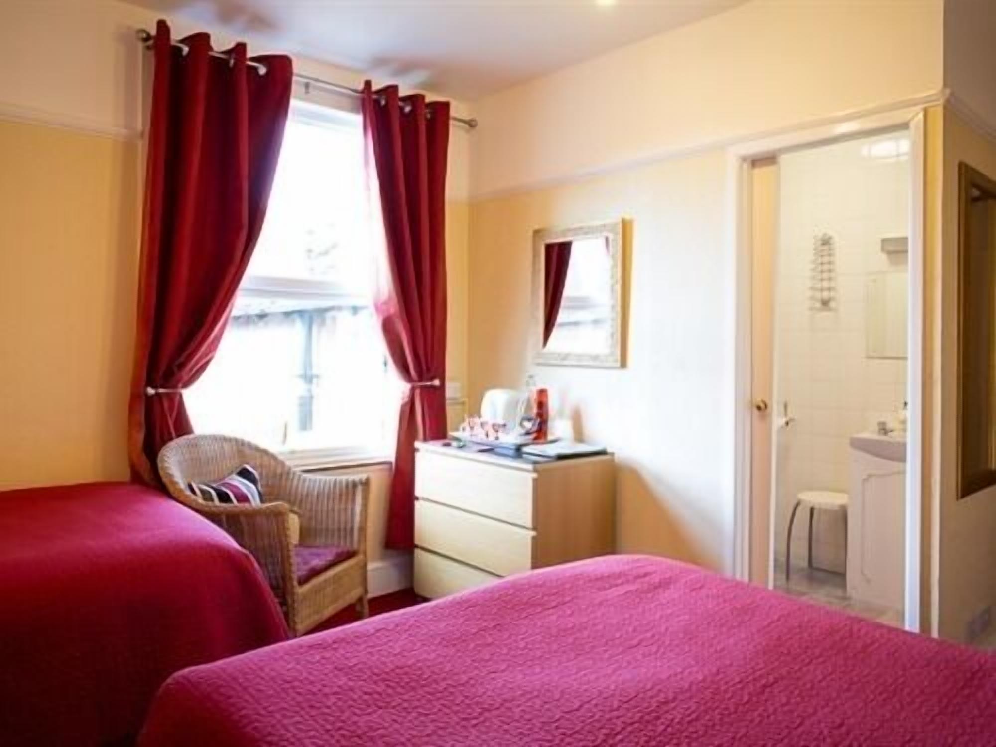 A hotel room with two beds, a desk, a chair, and a window with red curtains.