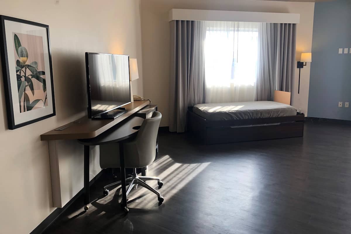 Habitación de hotel con una cama, un escritorio con computadora, una silla, un televisor y una ventana con cortinas.