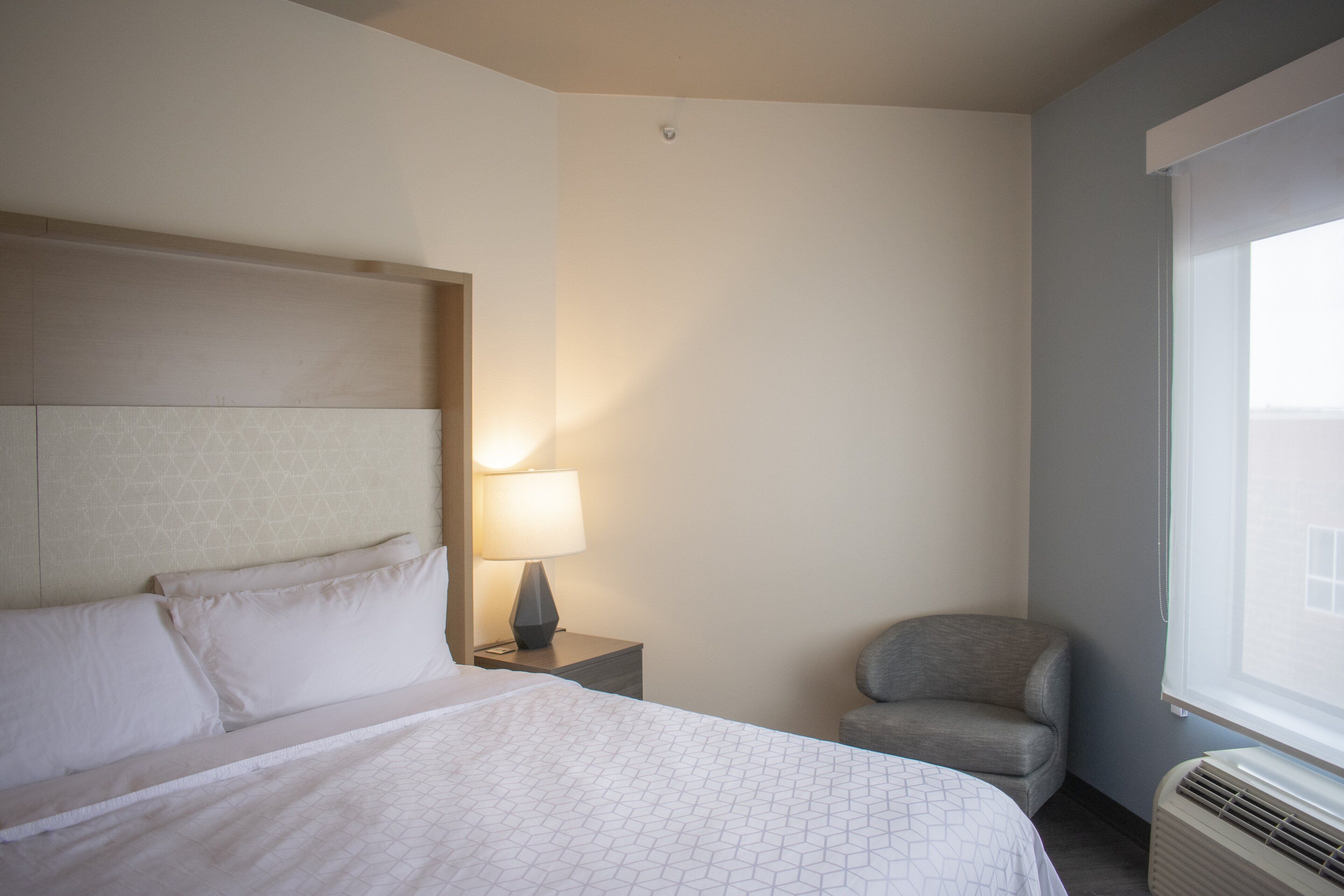 A hotel room with a bed, a chair, a nightstand with a lamp, and a window.