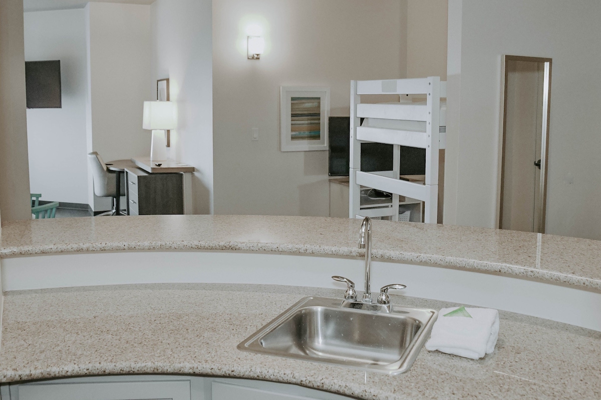 A kitchen with a stainless steel sink, a countertop, and a mirror reflecting the room.