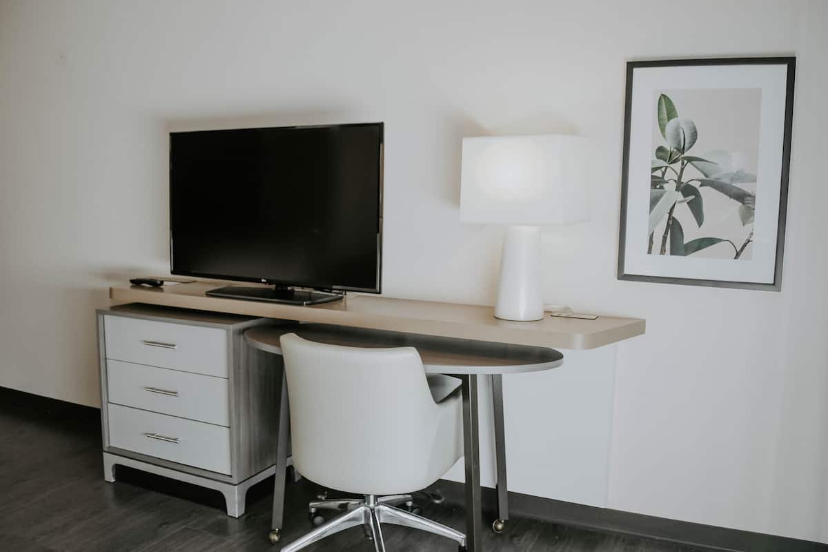 Una habitación de hotel moderna con escritorio, televisor de pantalla plana, una silla blanca, lámpara de escritorio y una planta enmarcada en la pared.