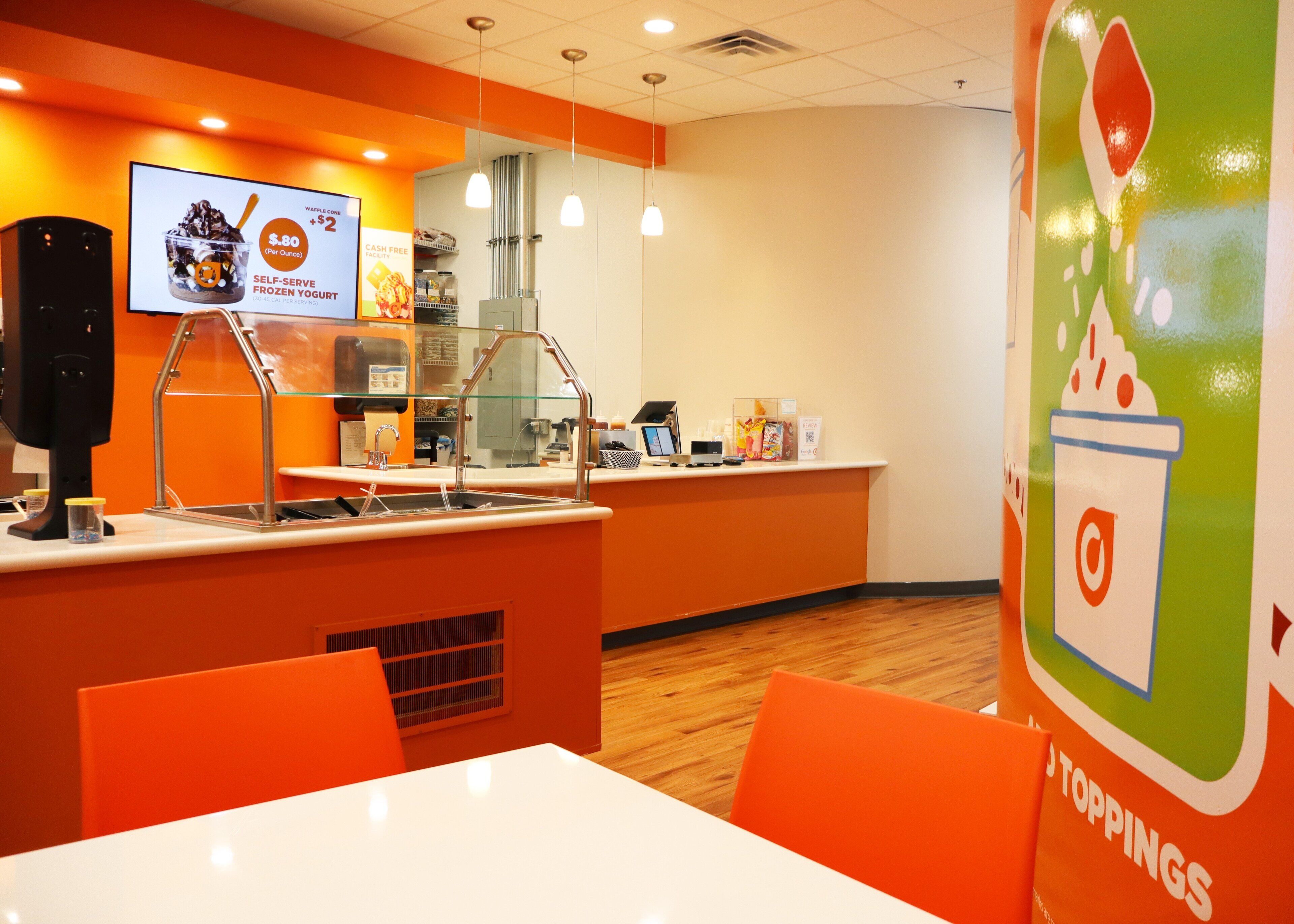 Un espace proposant des yaourts glacés en libre-service, avec un système de paiement sans espèces, un menu numérique et un coin salon doté de chaises orange et d’une table blanche.