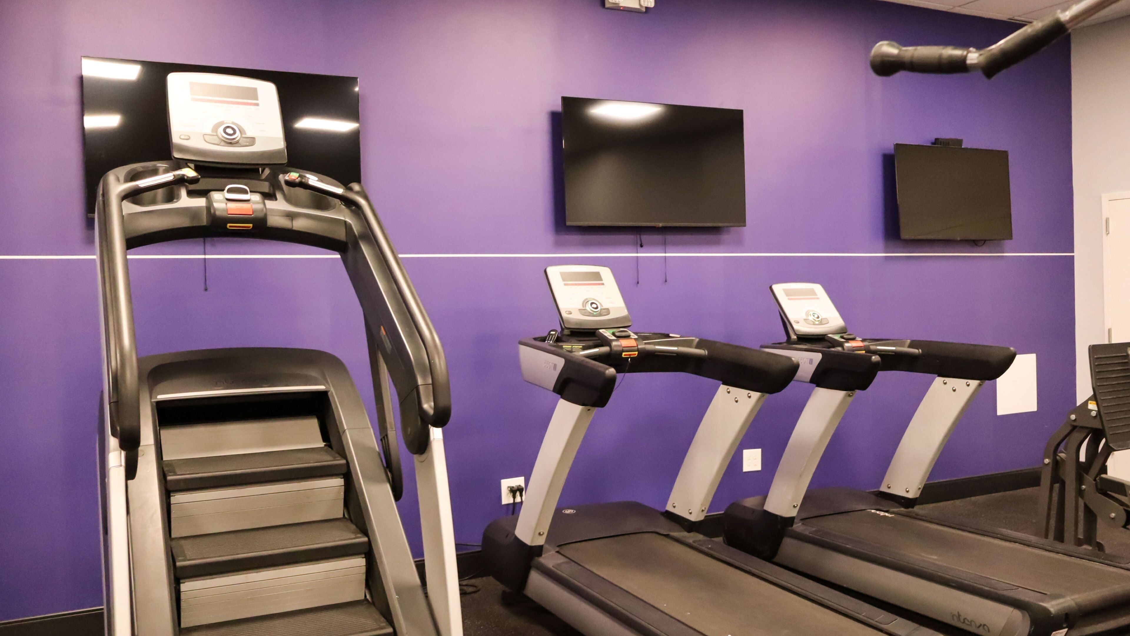 Une salle de sport avec des tapis de course et un appareil d’escalier, le tout adossé à un mur violet, avec un téléviseur fixé au-dessus.