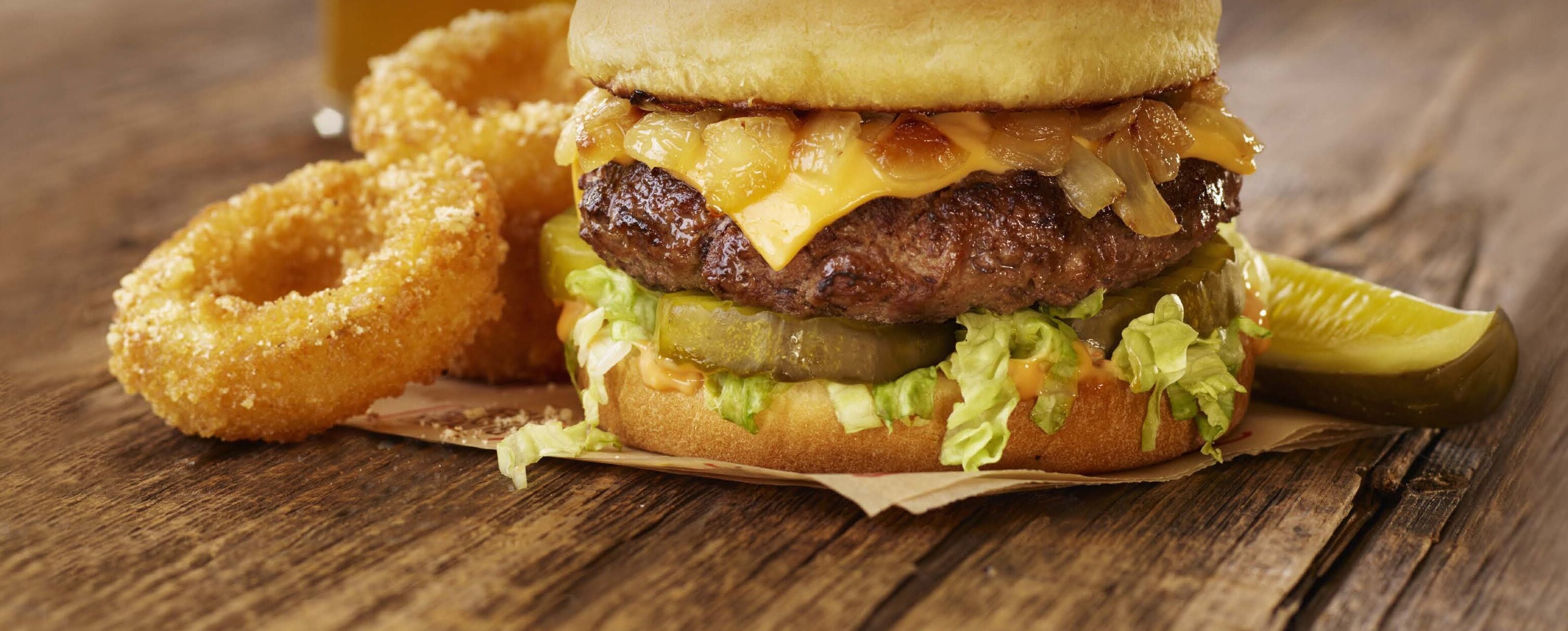 A burger with cheese, lettuce, and a pickle on a bun, accompanied by onion rings and a pickle spear on a wooden surface.