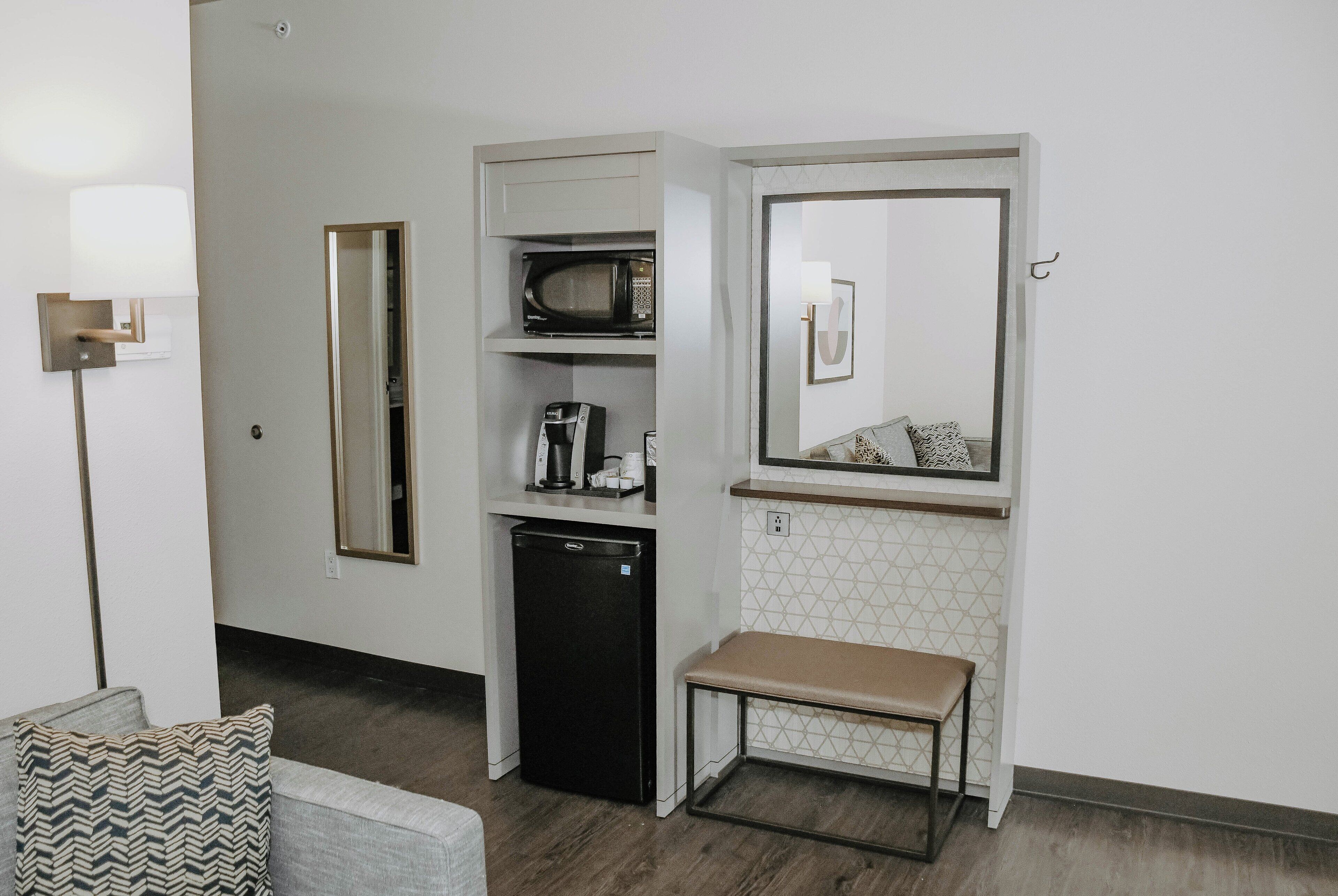A compact kitchenette with a microwave, coffee maker, and refrigerator, a bench, and a mirror above the counter.