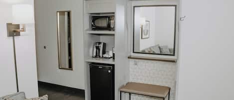 A compact kitchenette with a microwave, coffee maker, and refrigerator, a bench, and a mirror above the counter.
