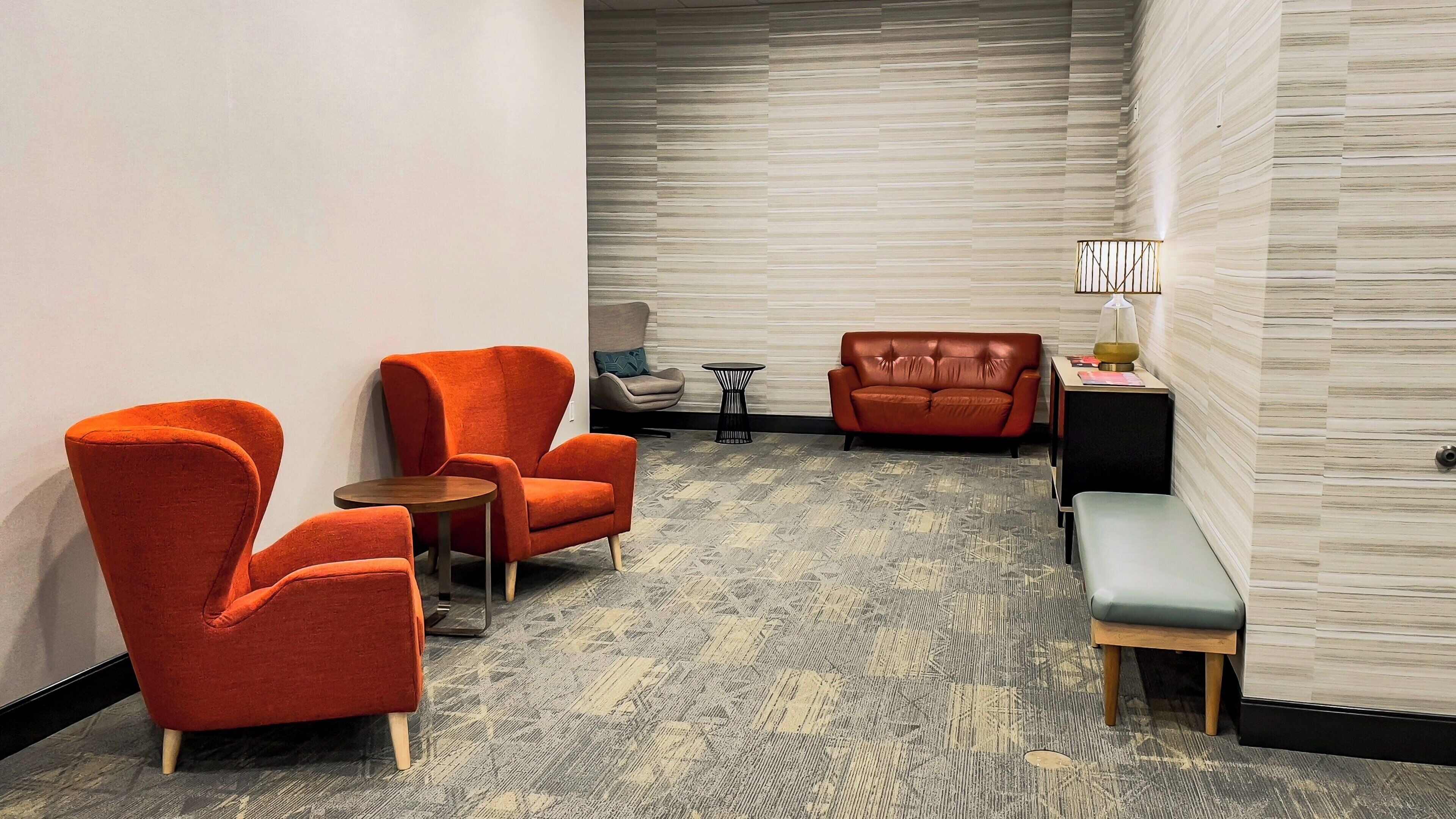 A hotel room with a seating area featuring two orange armchairs, a small round table, a sofa, and a bench. The room has a patterned carpet and a textured wall.