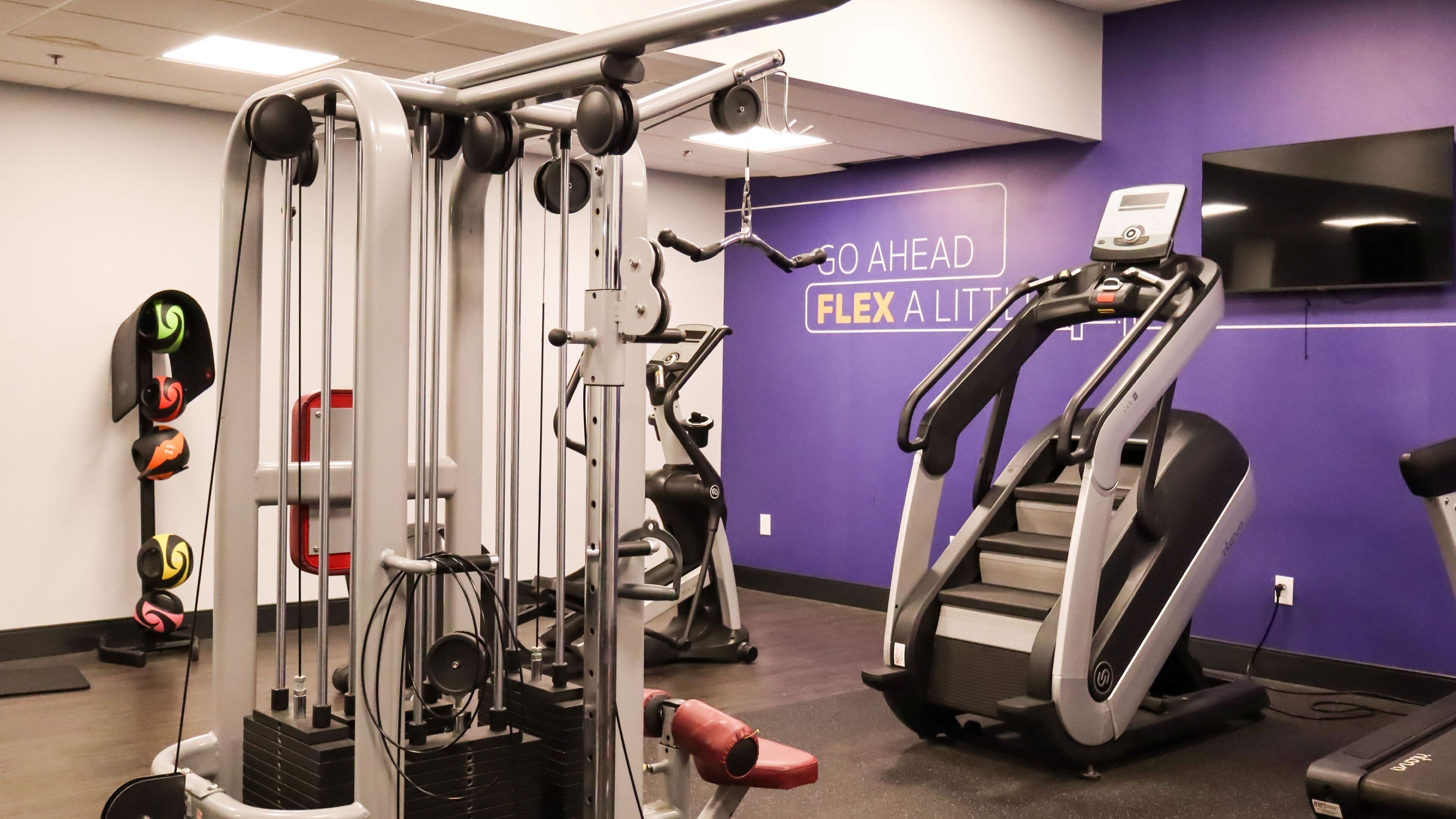 Un gimnasio con equipo de ejercicio, una cinta de correr y una pared morada con texto motivacional.