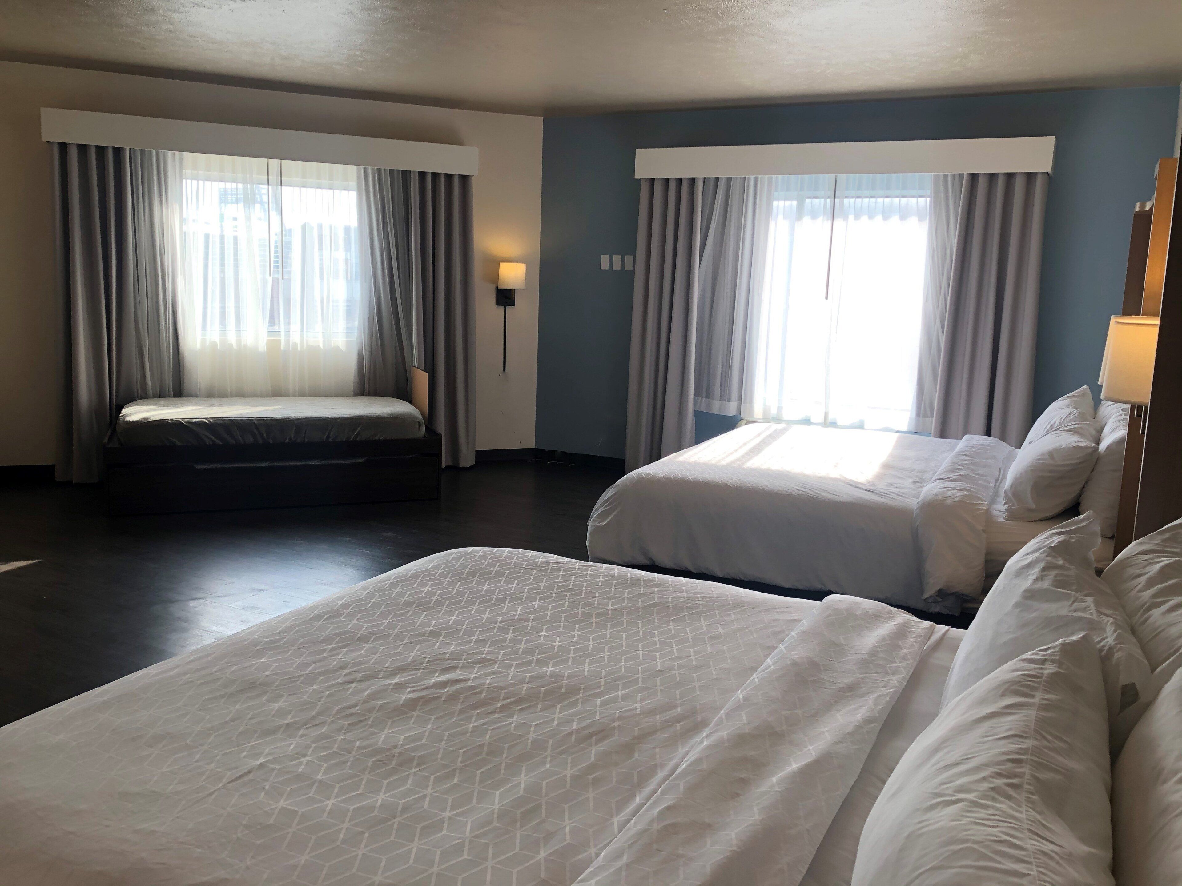 Habitación de hotel con dos camas, un ventanal con cortinas, una lámpara fijada a la pared y un suelo de madera oscura.