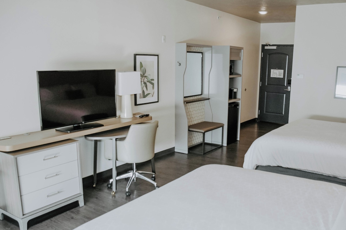 A hotel room with a desk, chair, TV, and two beds.