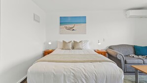 A bedroom with a bed, a chair, a small table, and a painting of a beach scene.