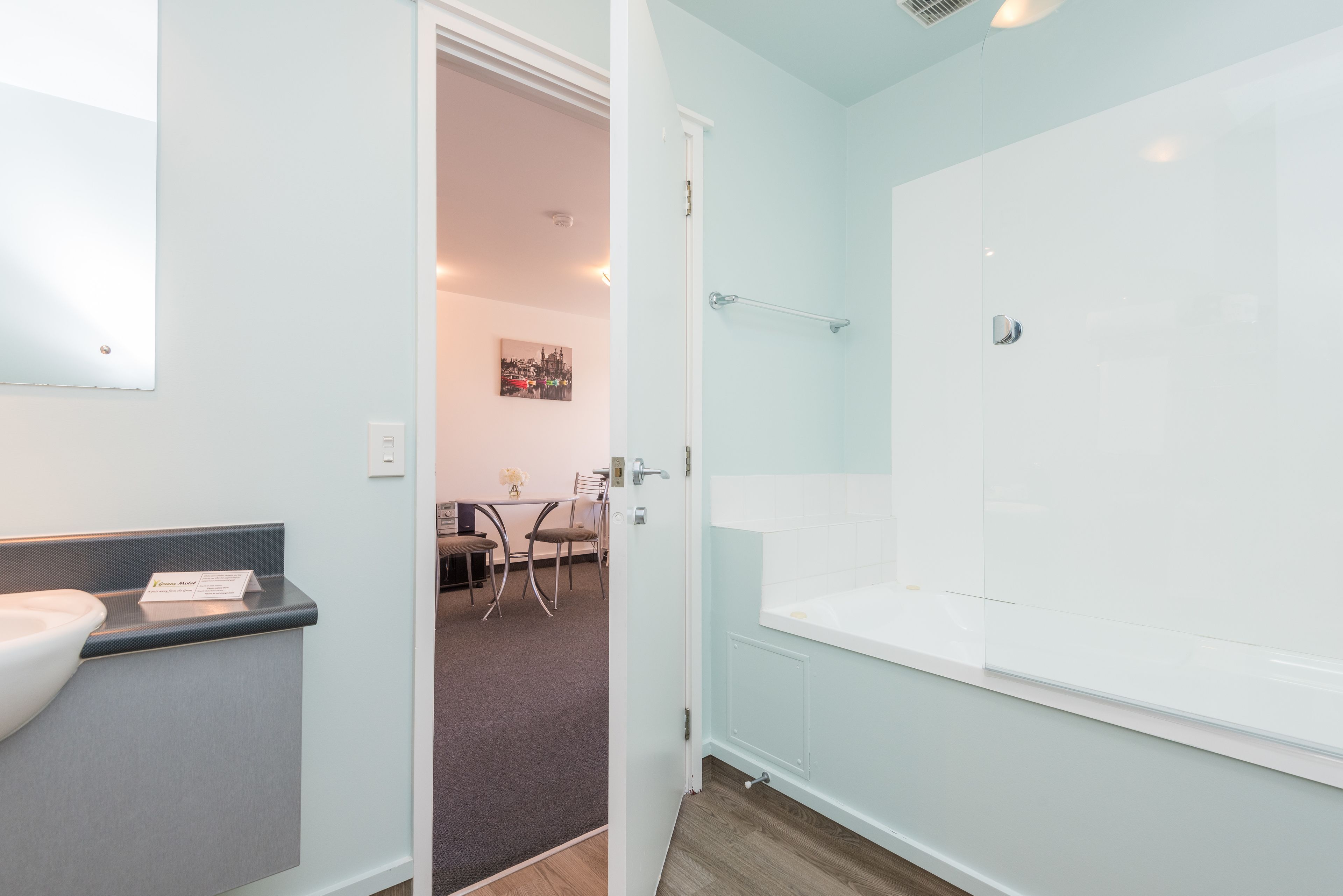 Un baño moderno con una bañera grande, un lavamanos y un espejo. Hay una puerta que da a un comedor con mesa y sillas.