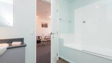 Un baño moderno con una bañera grande, un lavamanos y un espejo. Hay una puerta que da a un comedor con mesa y sillas.