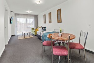 Una habitación de hotel moderna con comedor, una cama con cubrecama azul y blanco, una mesita con sillas y un ventanal con cortinas.
