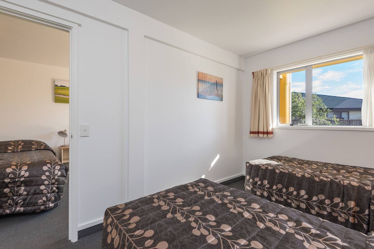 A room with two beds, a window with curtains, a door, and a painting on the wall.