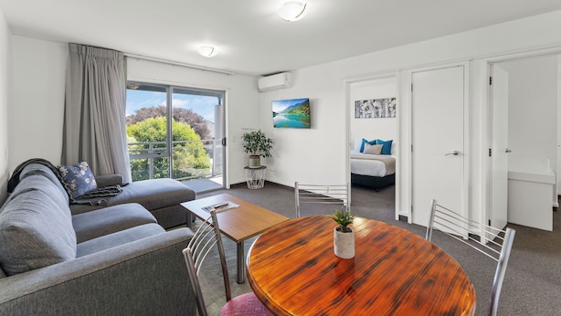 A modern living room with a grey sofa, a wooden dining table, and a large window offering a view of greenery.