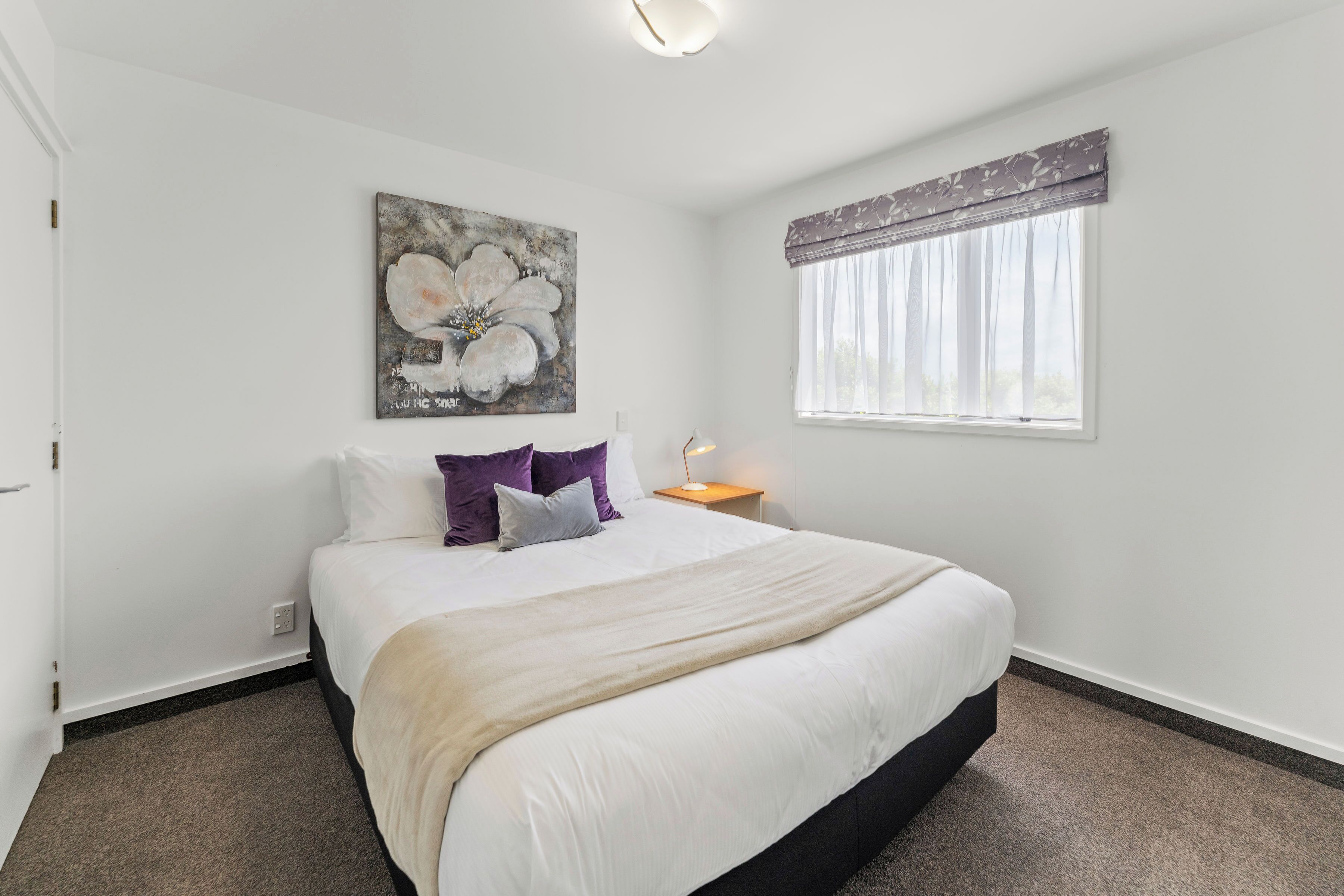 A bedroom with a large bed, a nightstand, a wall-mounted painting, and a window with curtains.