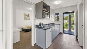 A compact kitchen with a microwave, coffee maker, and a small sink. There is a sofa in the adjacent living area and a door leading to a balcony with a view of greenery.