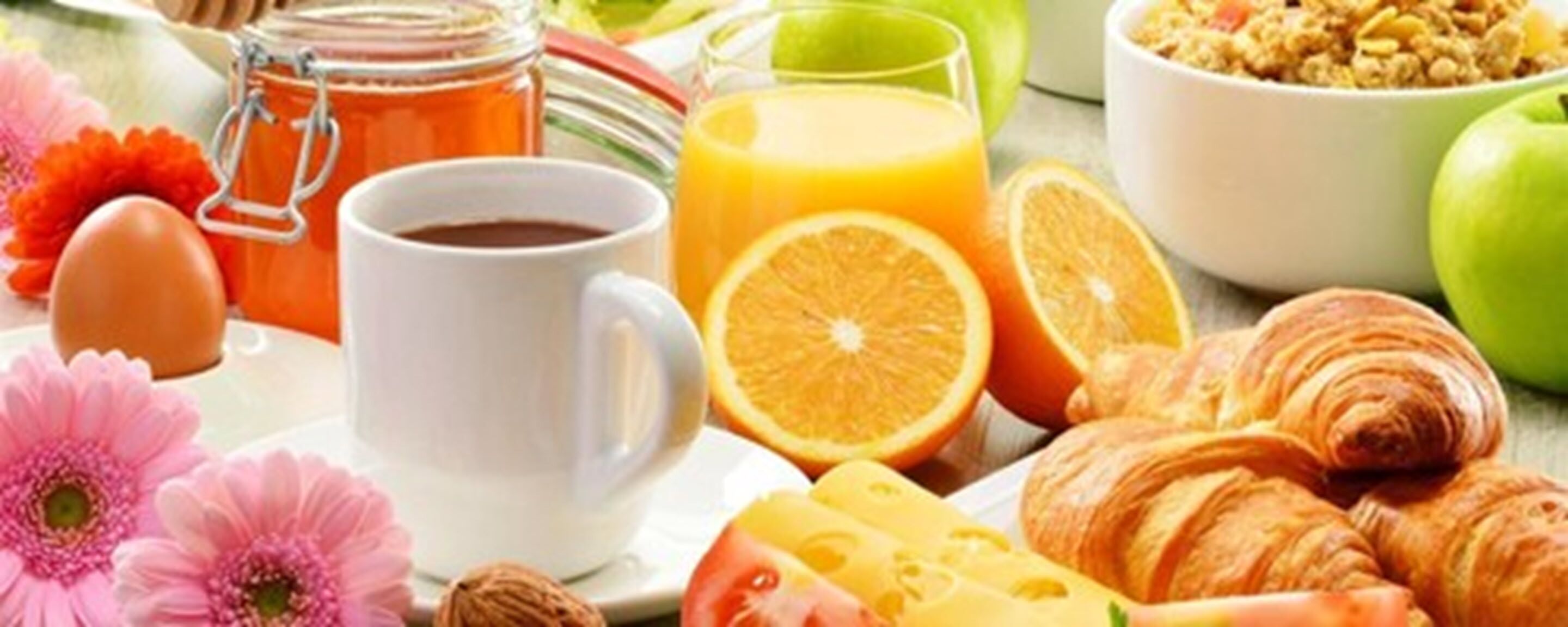 Una mesa con desayuno: una taza de café, jugo de naranja, croissants, queso y flores.