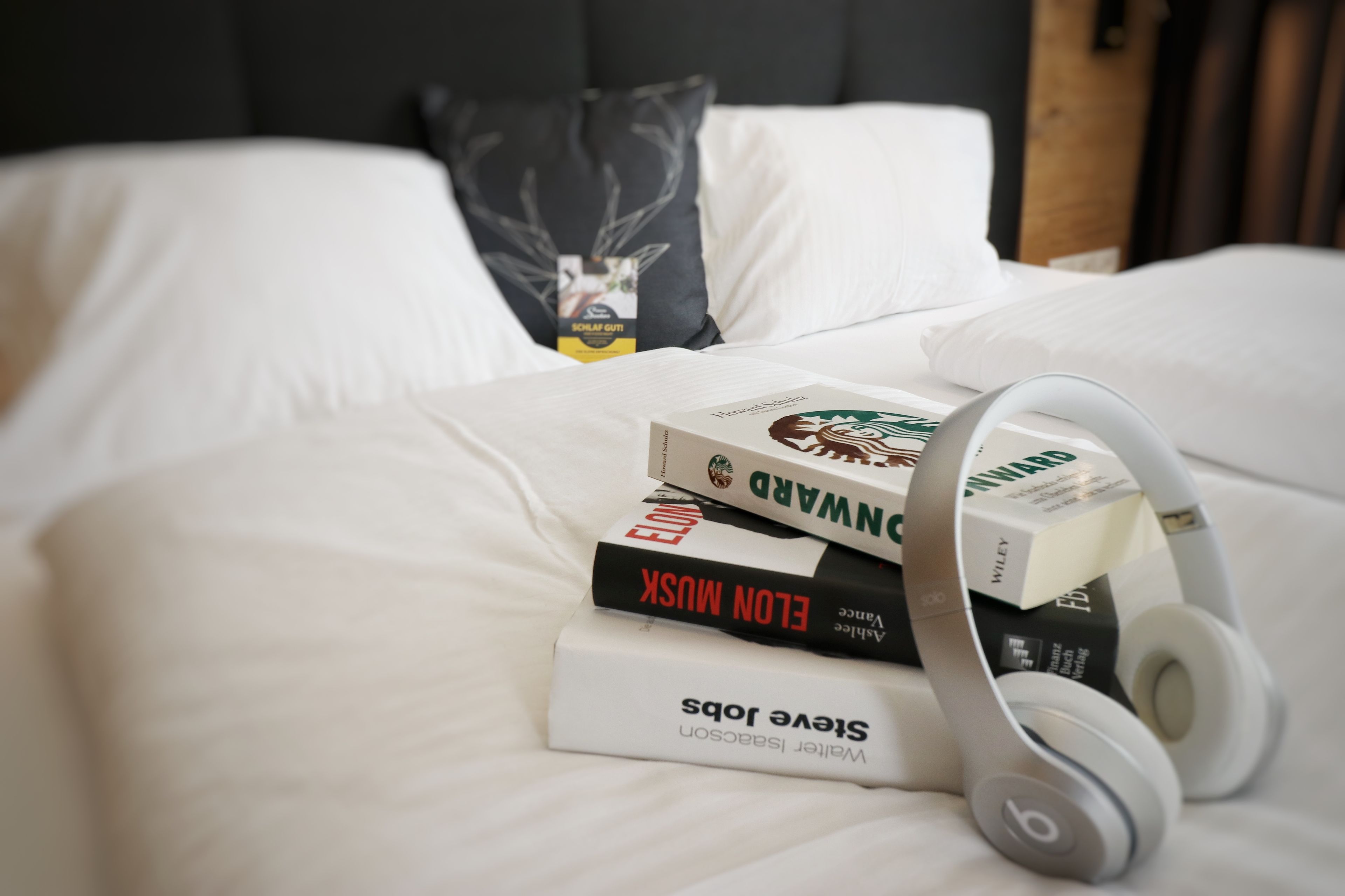 Una cama bien hecha con sábanas blancas, una almohada y una pila de libros, incluyendo títulos como 'Elon Musk' y 'Howard Stern'.