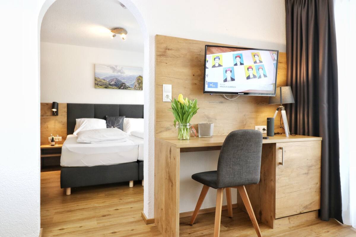 Una habitación de hotel con una cama, un escritorio con una silla, un televisor y una pequeña mesa con un jarrón de flores.