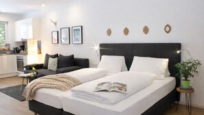 Une chambre moderne avec un grand lit, un canapé, une petite table avec une plante, et un coin cuisine visible au fond.