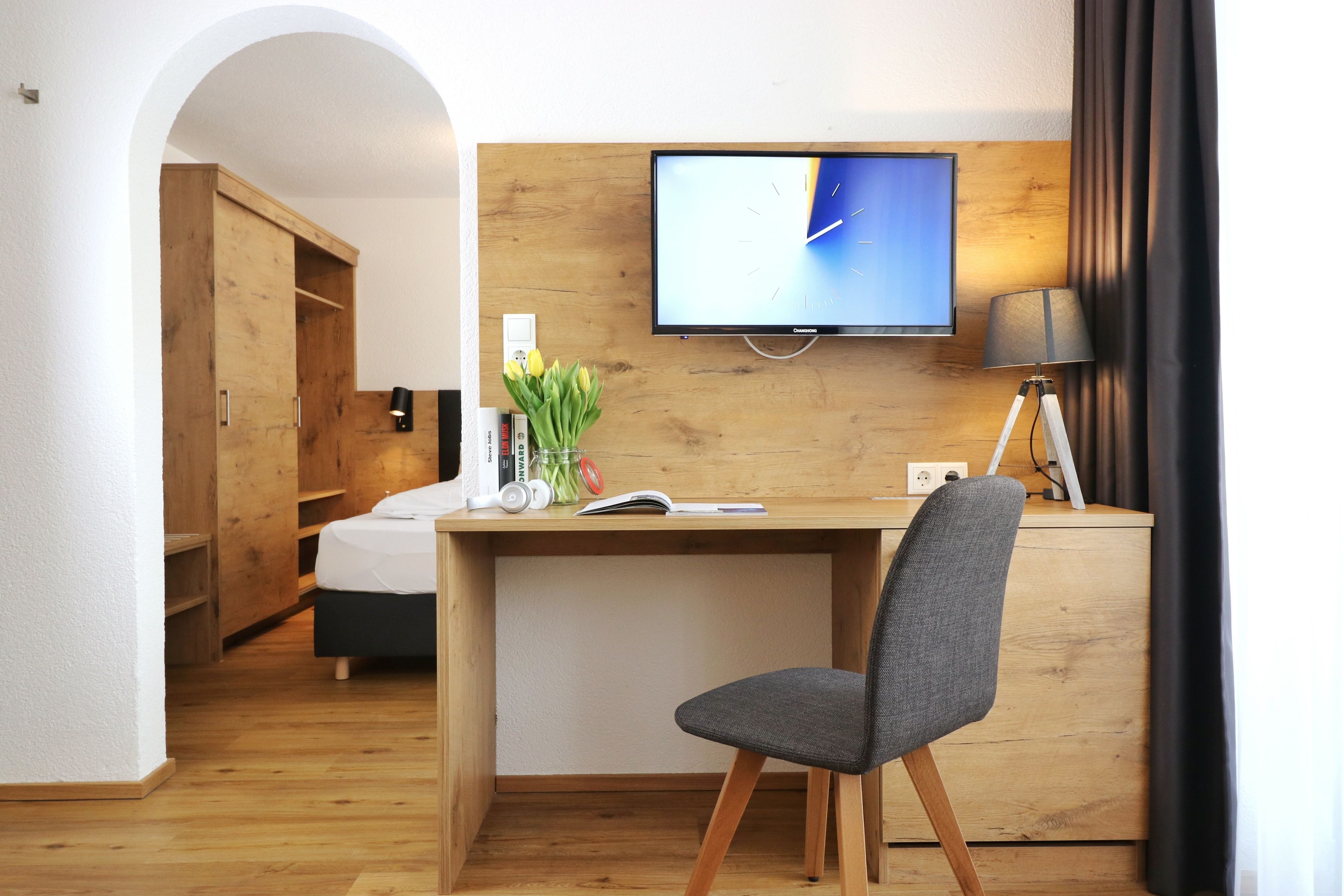 Una habitación de hotel moderna con un escritorio de madera, una silla gris, un televisor de pantalla plana, una lámpara y una estantería.