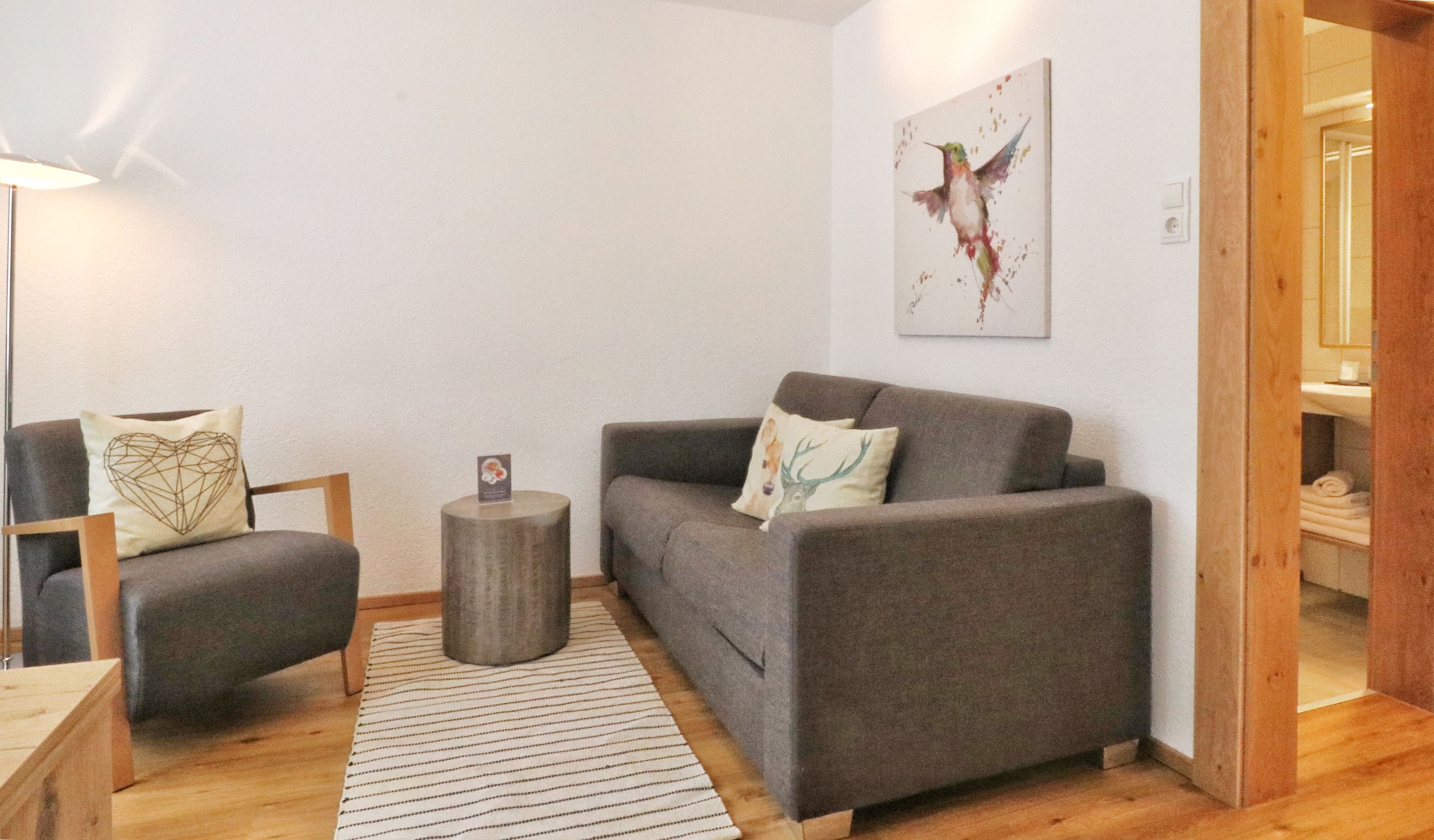 Un salon moderne avec un canapé gris, une table d’appoint en bois, un tapis à rayures et une œuvre d’art encadrée accrochée au mur.