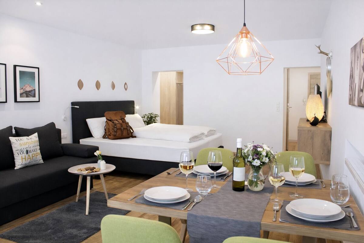Una habitación de hotel moderna con cama, sofá, una mesa de comedor para dos personas y una pequeña mesita de noche con un florero.