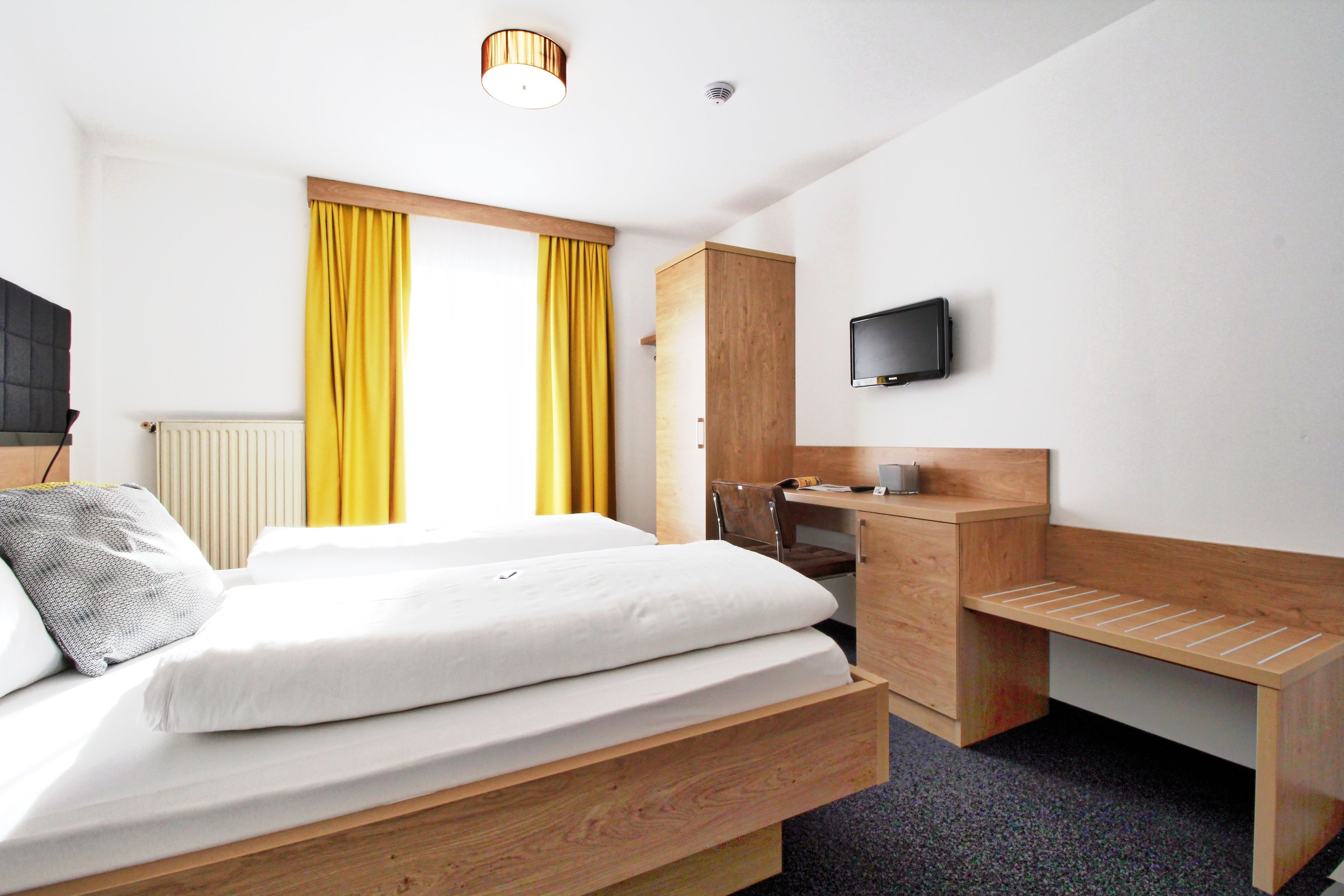 Habitación de hotel con dos camas, un escritorio de madera, televisión y ventana con cortinas amarillas.