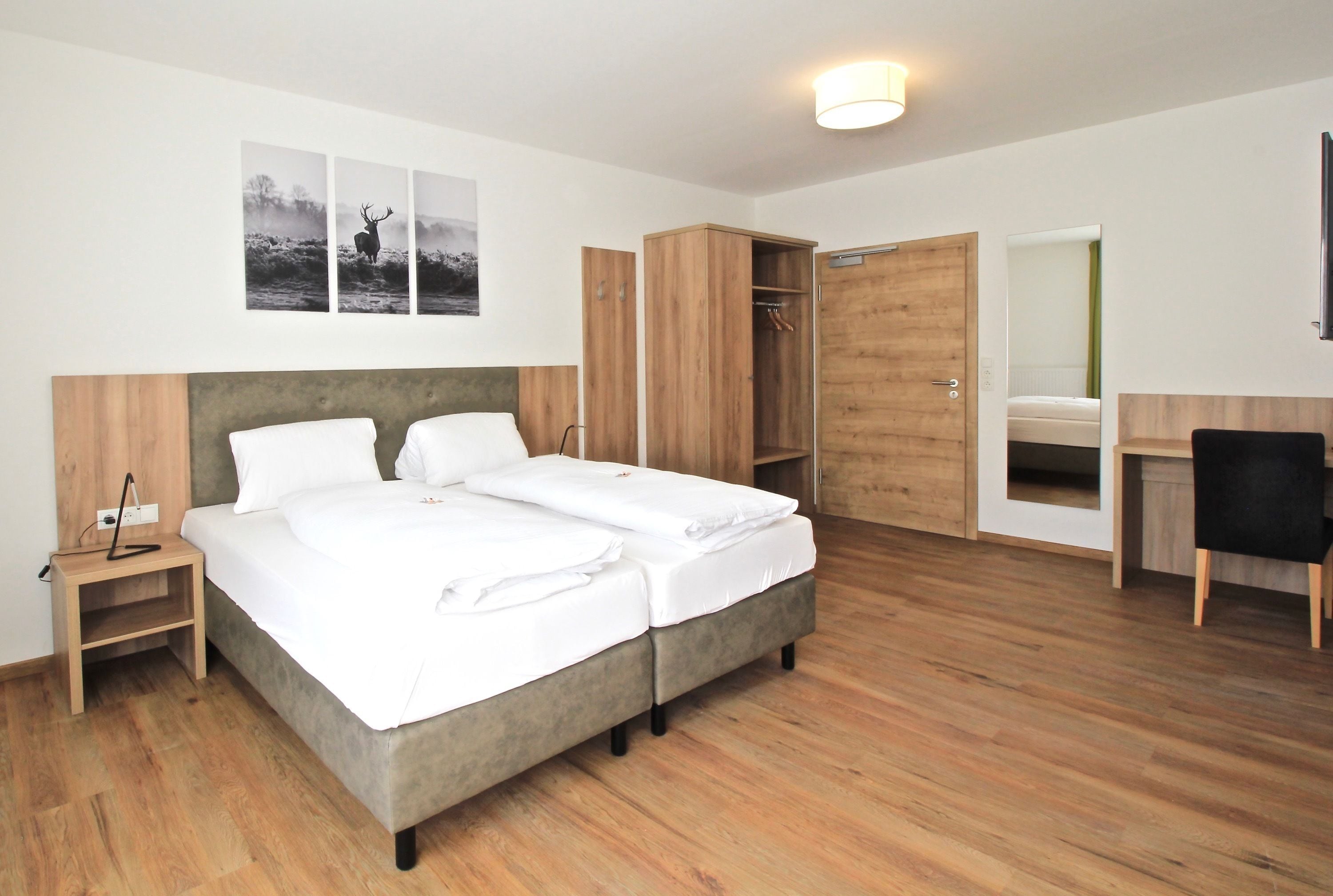 A modern hotel room with a double bed, wooden wardrobe, a desk with a chair, and a mirror.