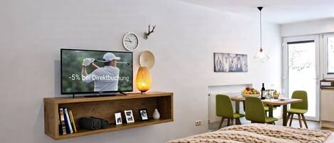 Quarto moderno com televisão, prateleira de madeira com livros, abajur e cama.