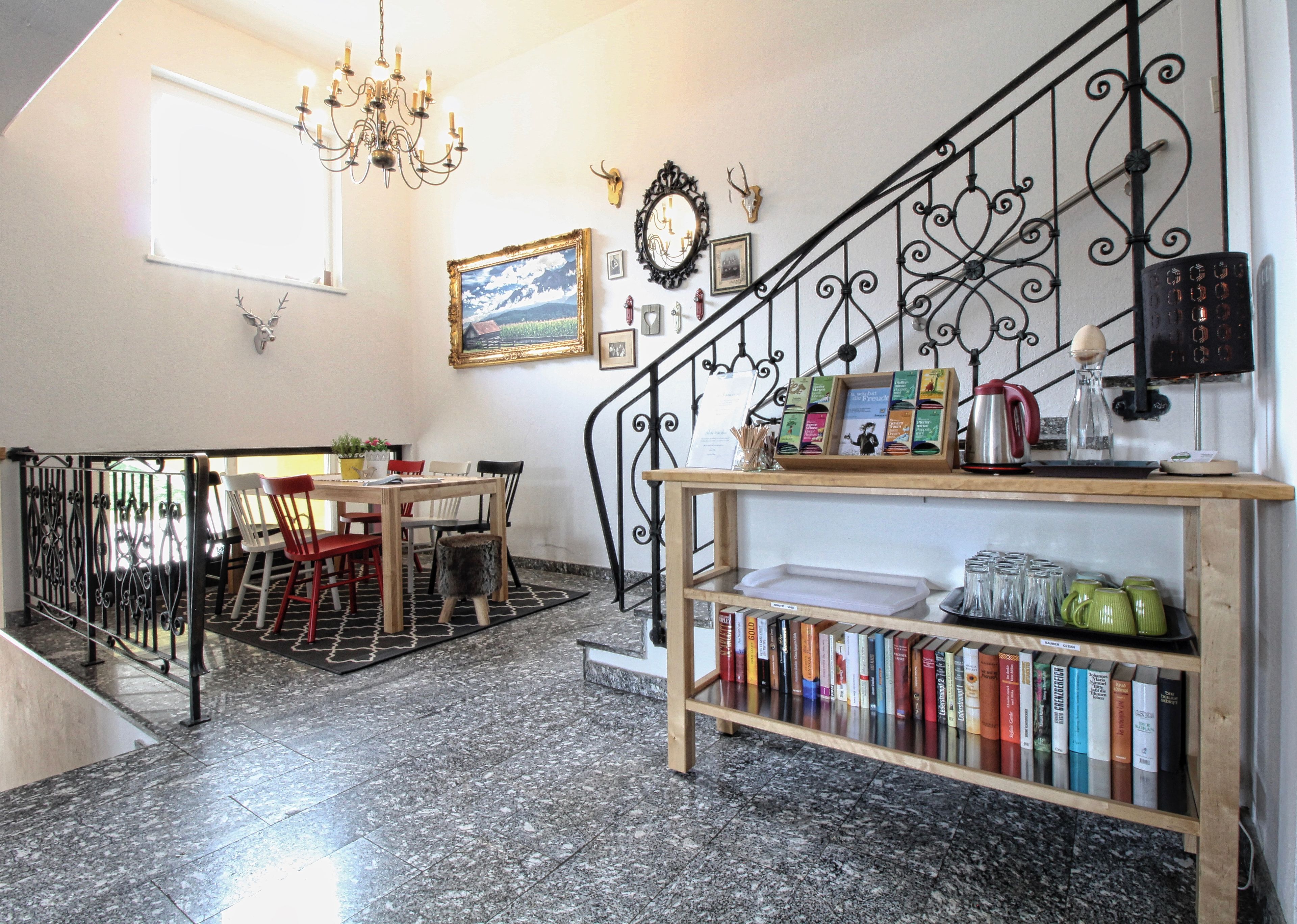 A dining area with a table and chairs, a bookshelf with books, a chandelier, and a staircase with a wrought iron railing.