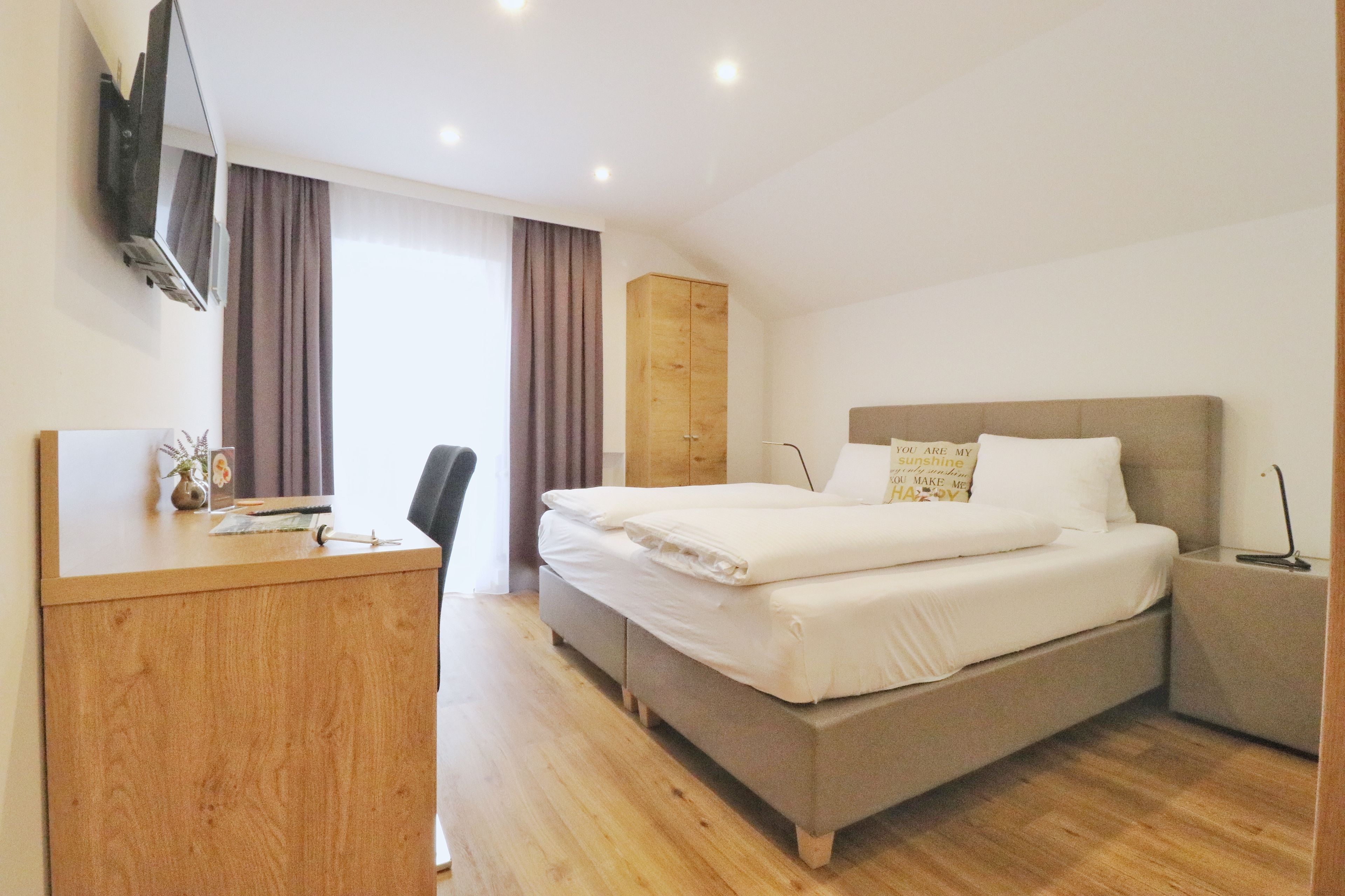 Ein modernes Hotelzimmer mit einem Holztisch, einem Bett mit Kopfteil, einem Nachttisch und einem an der Wand montierten Fernseher.