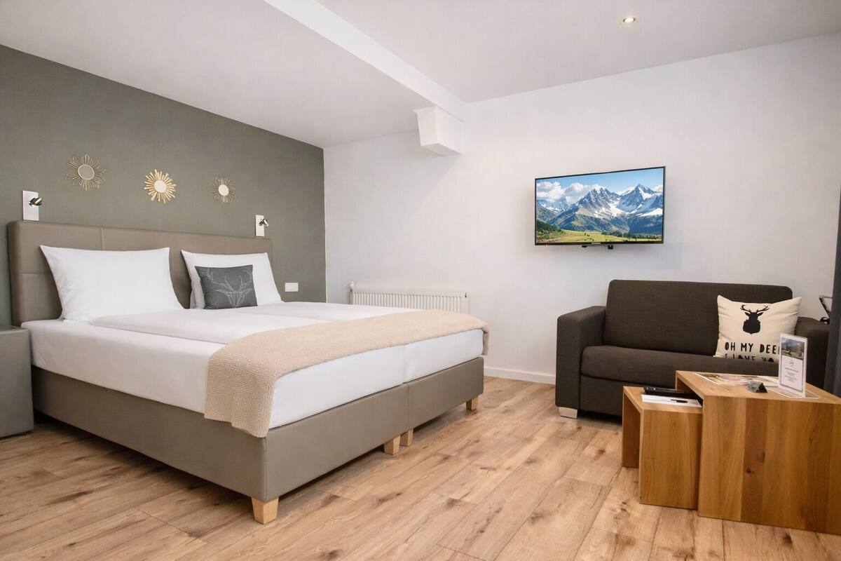 Un dormitorio moderno con cama, sofá, mesa de centro y un cuadro de paisaje montañoso en la pared.