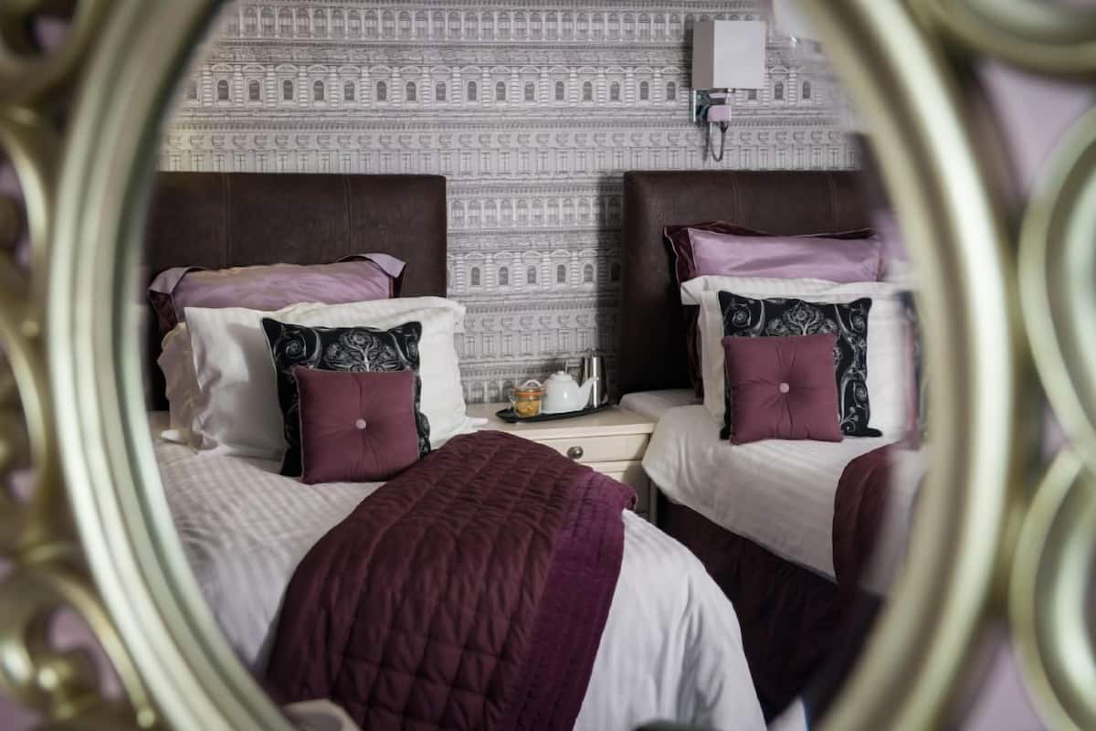 A hotel room with two beds, each with a purple headboard, a nightstand with a lamp, and a patterned wallpaper.