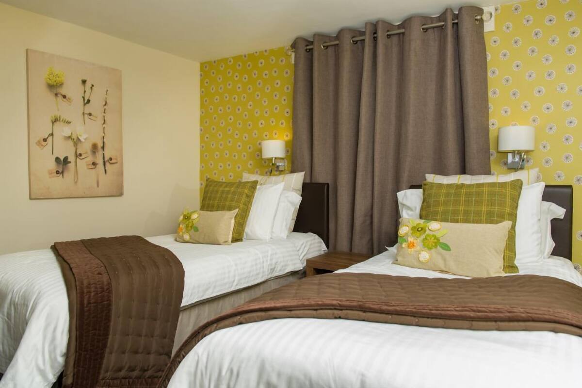 A hotel room with two beds, each with a brown blanket, white bedding, and decorative pillows. There are wall-mounted lamps and a floral artwork above the beds.