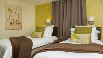 A hotel room with two beds, each with a brown blanket, white bedding, and decorative pillows. There are wall-mounted lamps and a floral artwork above the beds.
