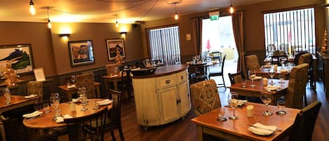 Un restaurante con mesas y sillas de madera, luces colgantes, cuadros en las paredes y un amplio ventanal con persianas.